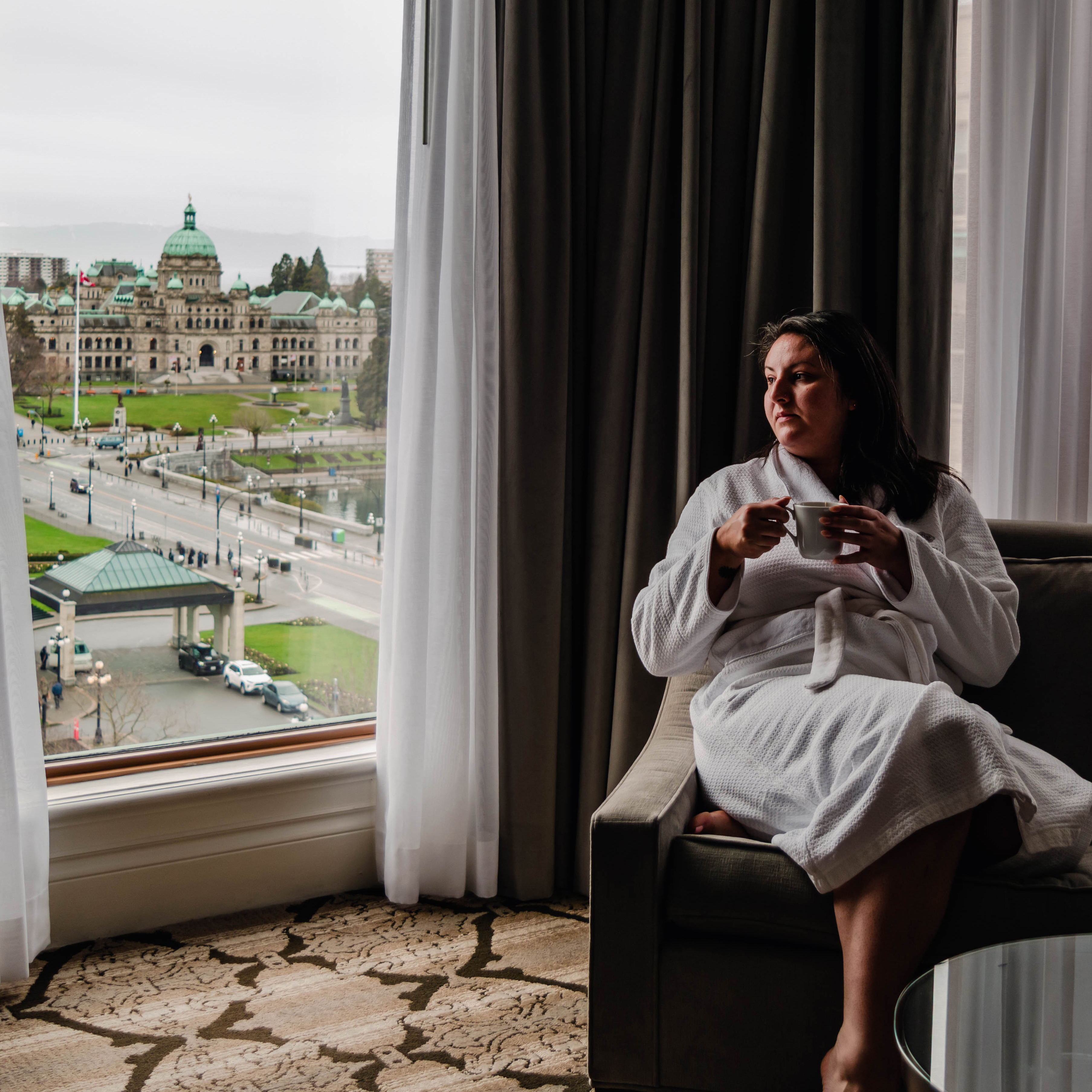 A woman relaxes on a couch with a cup of tea overlooking the BC Legislature Buildings in Victoria BC